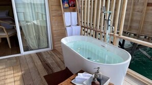Una habitación con una bañera grande, piso de madera, una mesita con una toalla y una botella, y vista al agua.