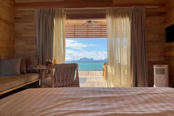 A wooden room with a bed, a chair, a small table with a bottle, and a view of the sea. - Sisipan Island Water Resort (Semporna)
