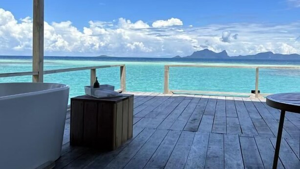 A wooden deck with a white bathtub, a small wooden table, and a view of the ocean and mountains. - Sisipan Island Water Resort (Semporna)
