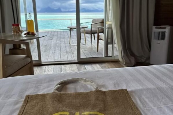 A hotel room with a bed, a table with a bottle and snacks, a chair, and a view of the ocean.