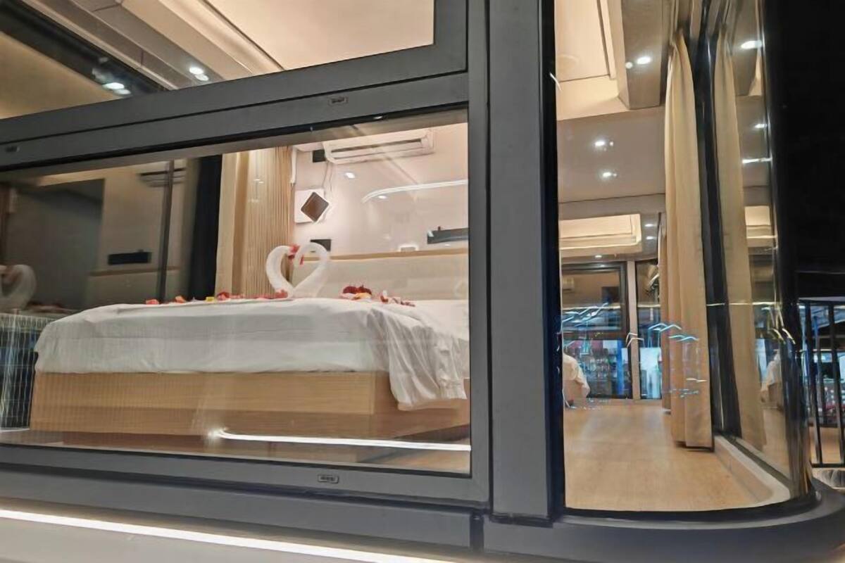 A modern hotel room with a large bed, a glass partition, and a view of the hotel's interior.