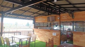 An outdoor restaurant with wooden tables and chairs, a counter with a menu board, and a view of the surrounding area.