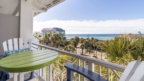 Ein Balkon mit einem grünen Tisch und weißen Stühlen, der auf einen Strand mit Palmen und ein Gebäude im Hintergrund blickt.