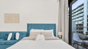 Ett modernt hotellrum med en stor säng, en blå stoppad soffa och en balkong med utsikt över en byggnad.