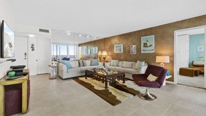 A spacious living room with a large sofa set, a coffee table, and a purple armchair. There are wall-mounted picture frames and a door leading to another room.