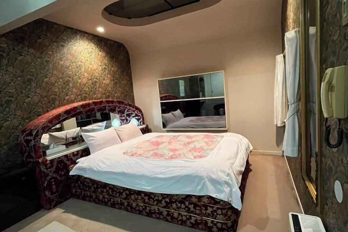 A bedroom with a curved headboard, a bed with a floral bedspread, a bedside table, a mirror, and a wall-mounted telephone.