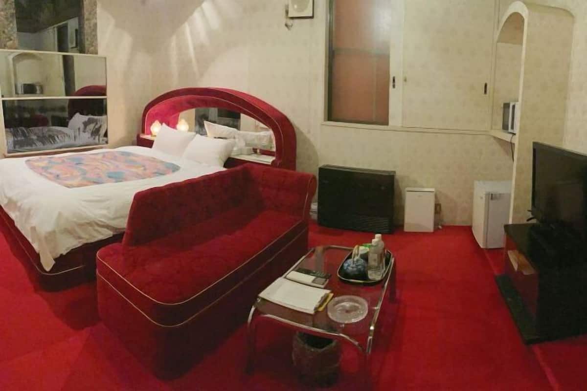 A hotel room with a bed, a red sofa, a small table with items, a TV, and a mirror.