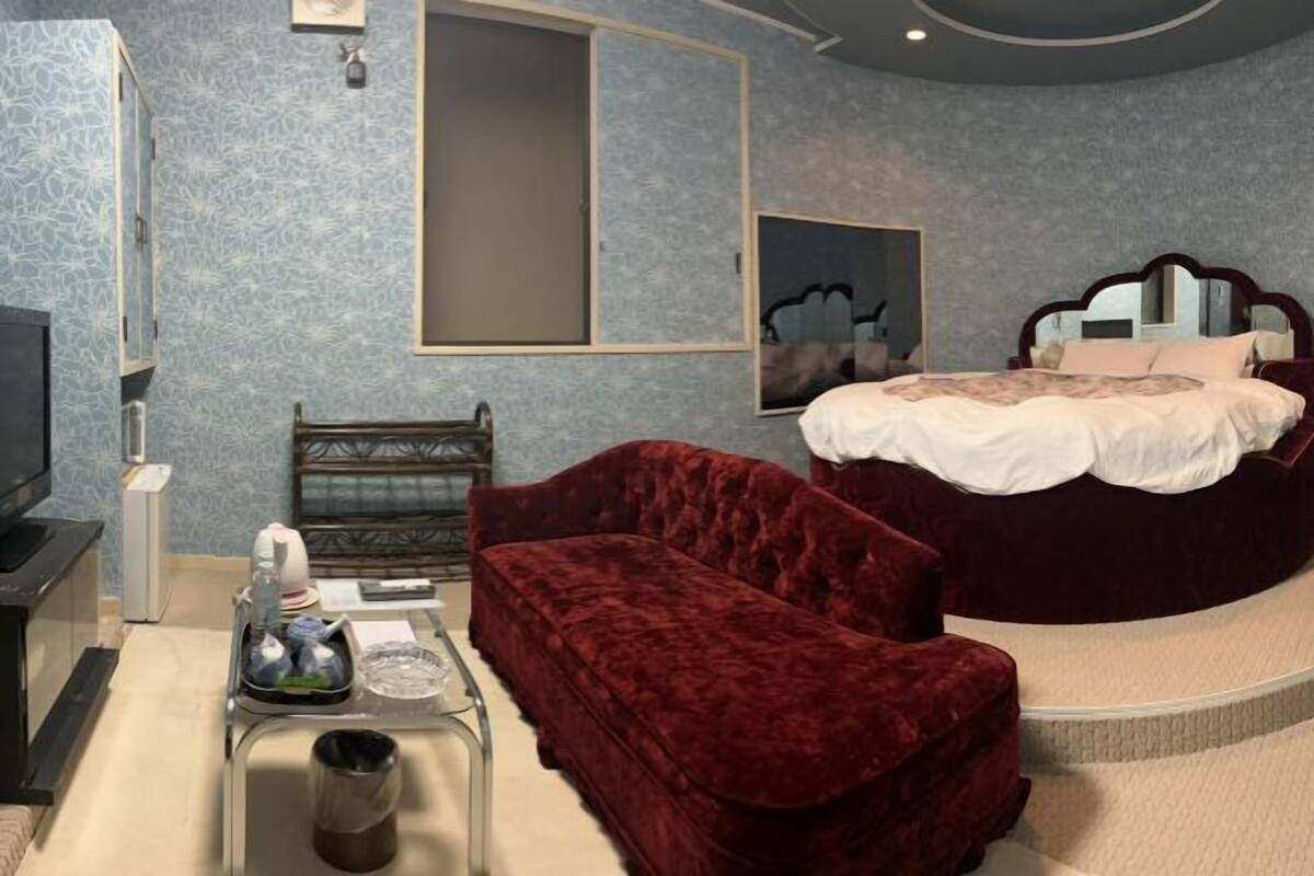 A bedroom with a bed, a red velvet chair, a small table with items, a TV, and a mirror.