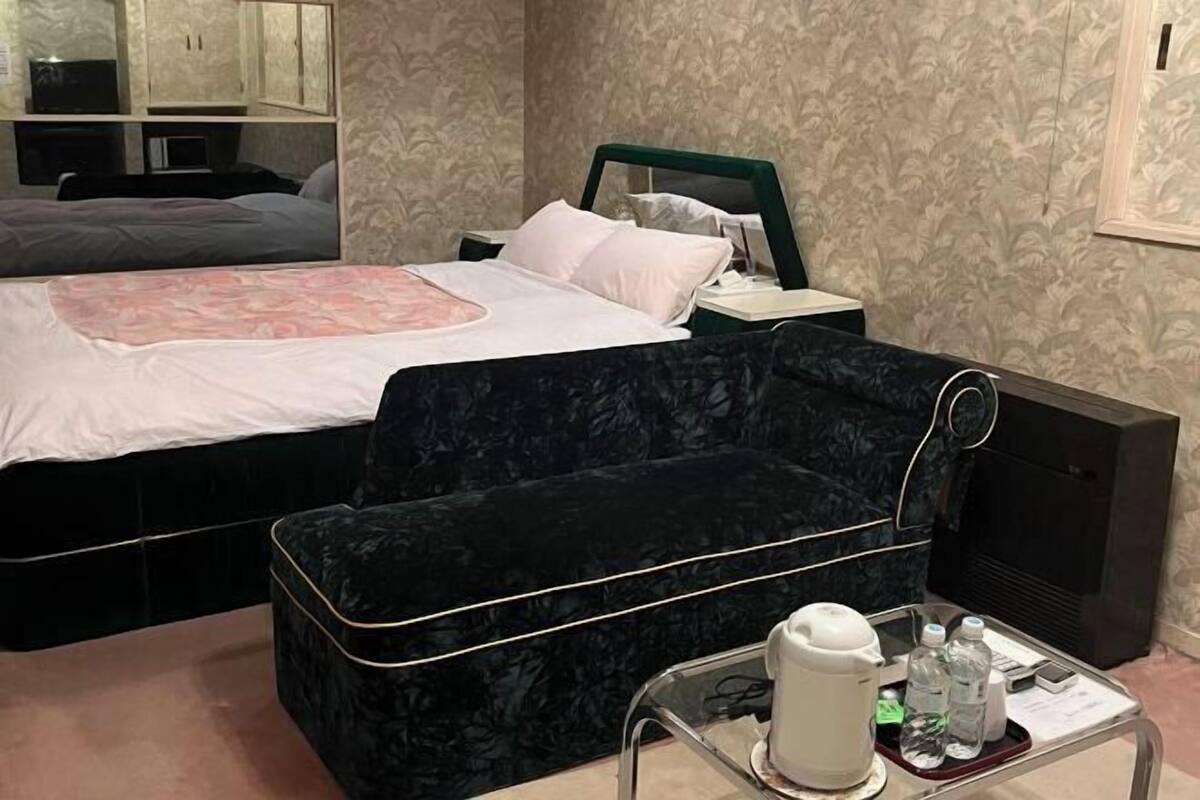 A hotel room with a bed, a black velvet sofa, a small table with a kettle and bottles, and a television.