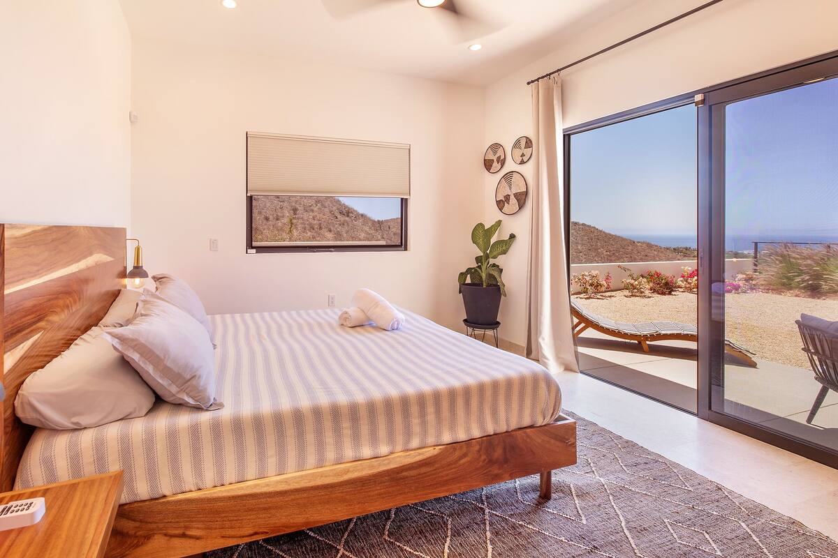 Une chambre avec un lit en bois, une lampe de chevet, une plante en pot et une vue sur la plage, visible à travers une porte-fenêtre coulissante.