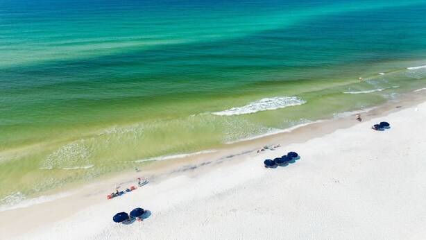 An aerial view of a beach with clear turquoise waters, a sandy shore, and a few umbrellas. - 30A Beachside - Steps to Beach! Centrally located, Pools and Tennis! (Santa Rosa Beach)