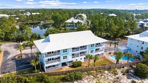 A multi-story hotel with balconies, a swimming pool, and a view of a lake and surrounding trees. - 30A Beachside - Steps to Beach! Centrally located, Pools and Tennis! (Santa Rosa Beach)