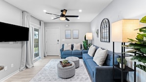 A modern living room with a blue sofa, ottomans, a flat-screen TV, and a ceiling fan. - 30A Beachside - Steps to Beach! Centrally located, Pools and Tennis! (Santa Rosa Beach)