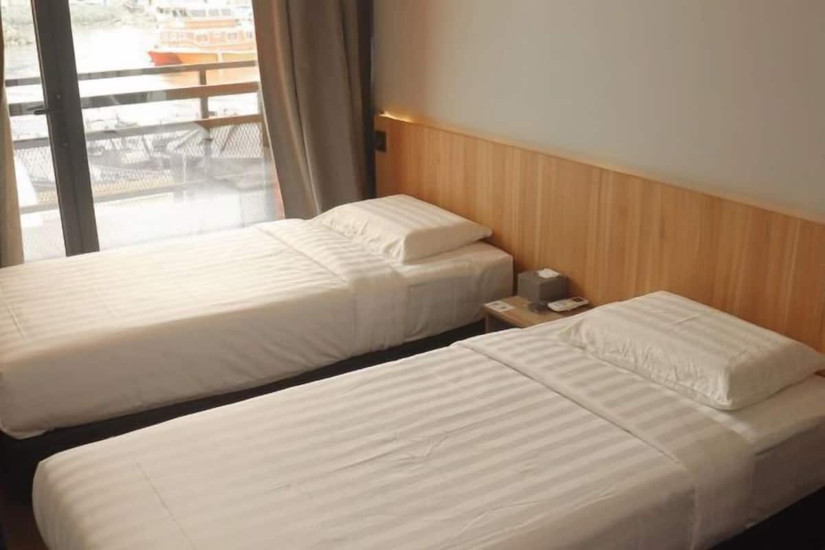 A hotel room with two beds, a wooden headboard, a nightstand with a lamp, and a view of a boat through a window.