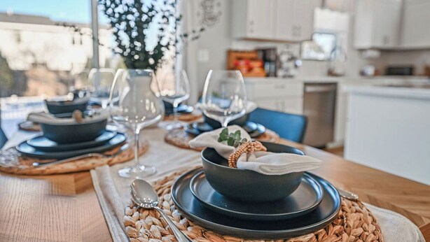 Una mesa de comedor dispuesta con platos, tazones y vasos, con una cocina al fondo.