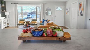 Una tabla de madera con varios quesos, frutas y embutidos en un moderno salón con vistas al exterior.
