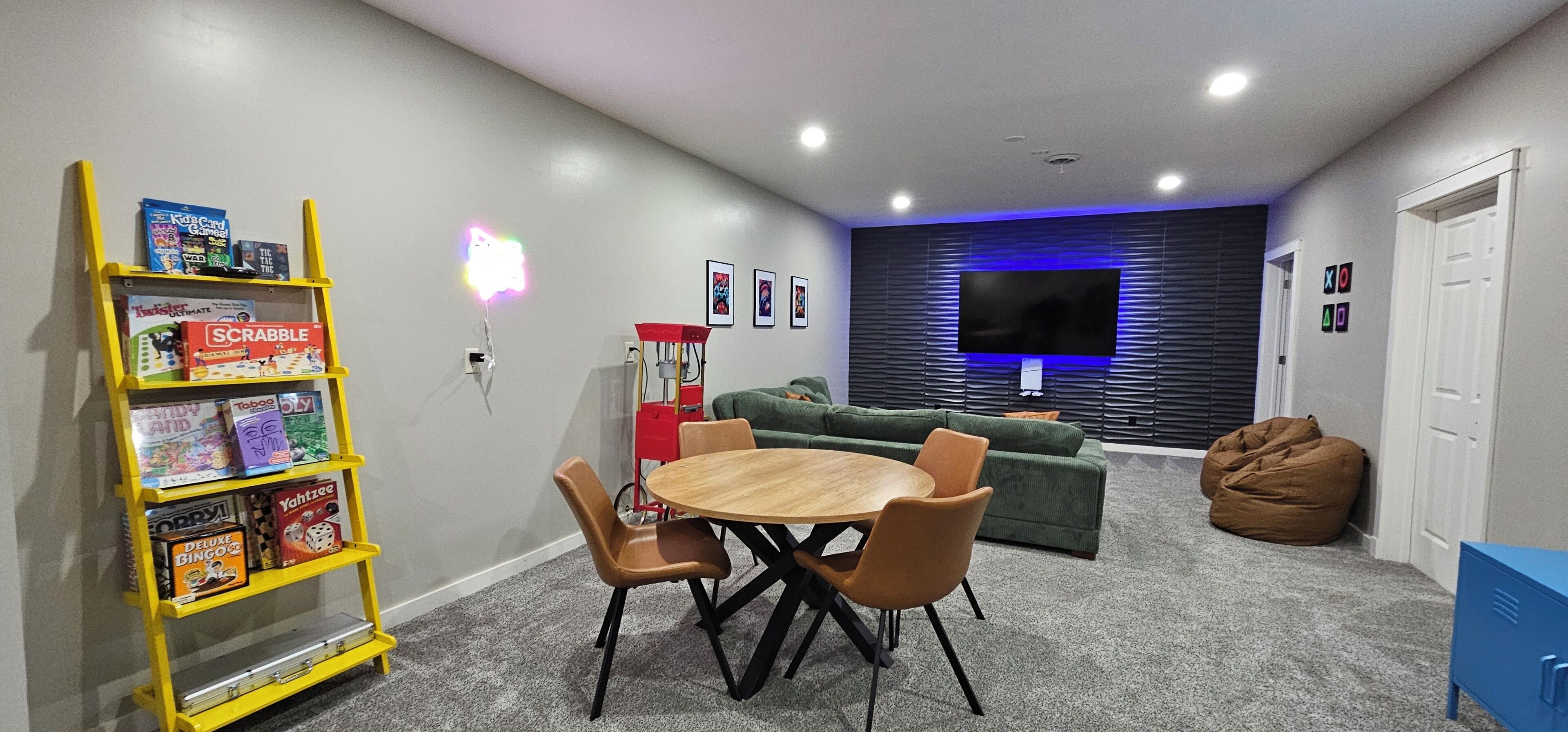 A room with a wooden table and chairs, a green sofa, a bean bag, a TV wall-mounted above a sofa, and a shelf with board games.