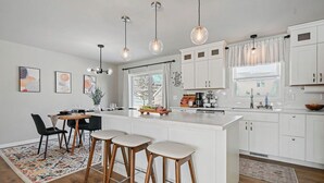 Una cocina moderna con armarios blancos, una isla central con taburetes, iluminación colgante y vistas al exterior a través de amplias ventanas.