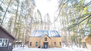 Puinen mökki, jonka päällä on metallikatto, ja jonka ympärillä kohoaa korkeita mäntyjä lumisessa maisemassa.