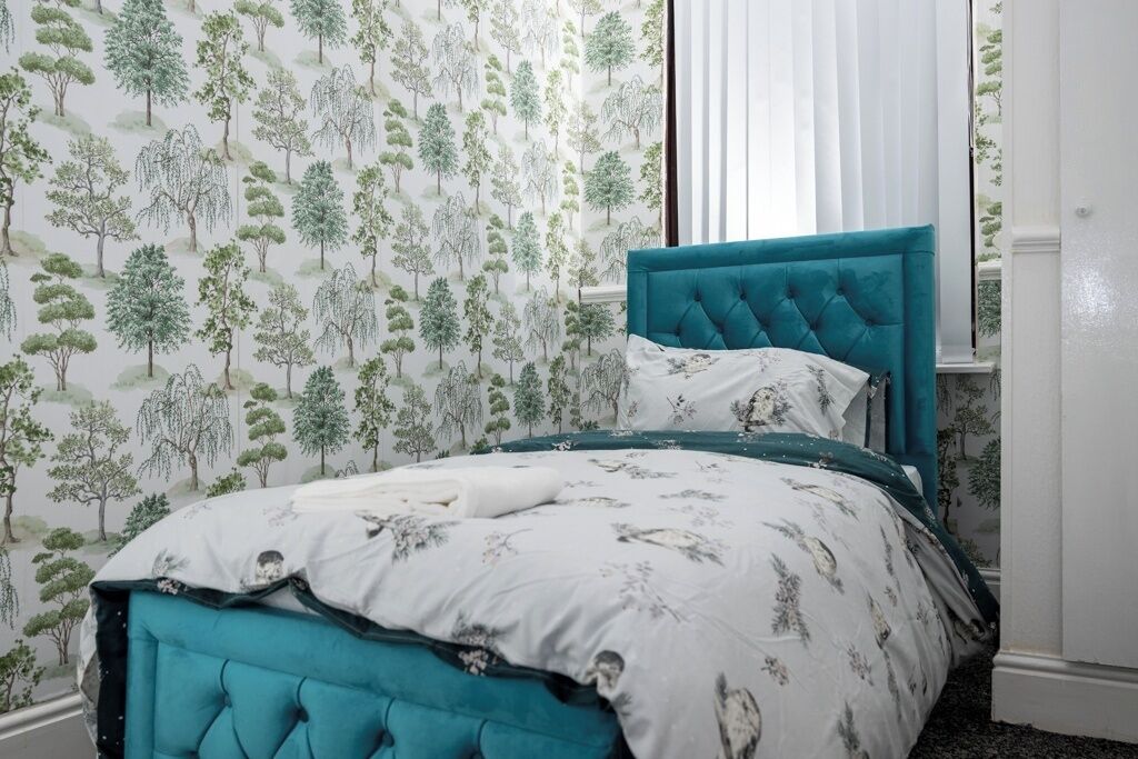A bedroom with a bed, a window with blinds, and a wall with a tree pattern wallpaper.
