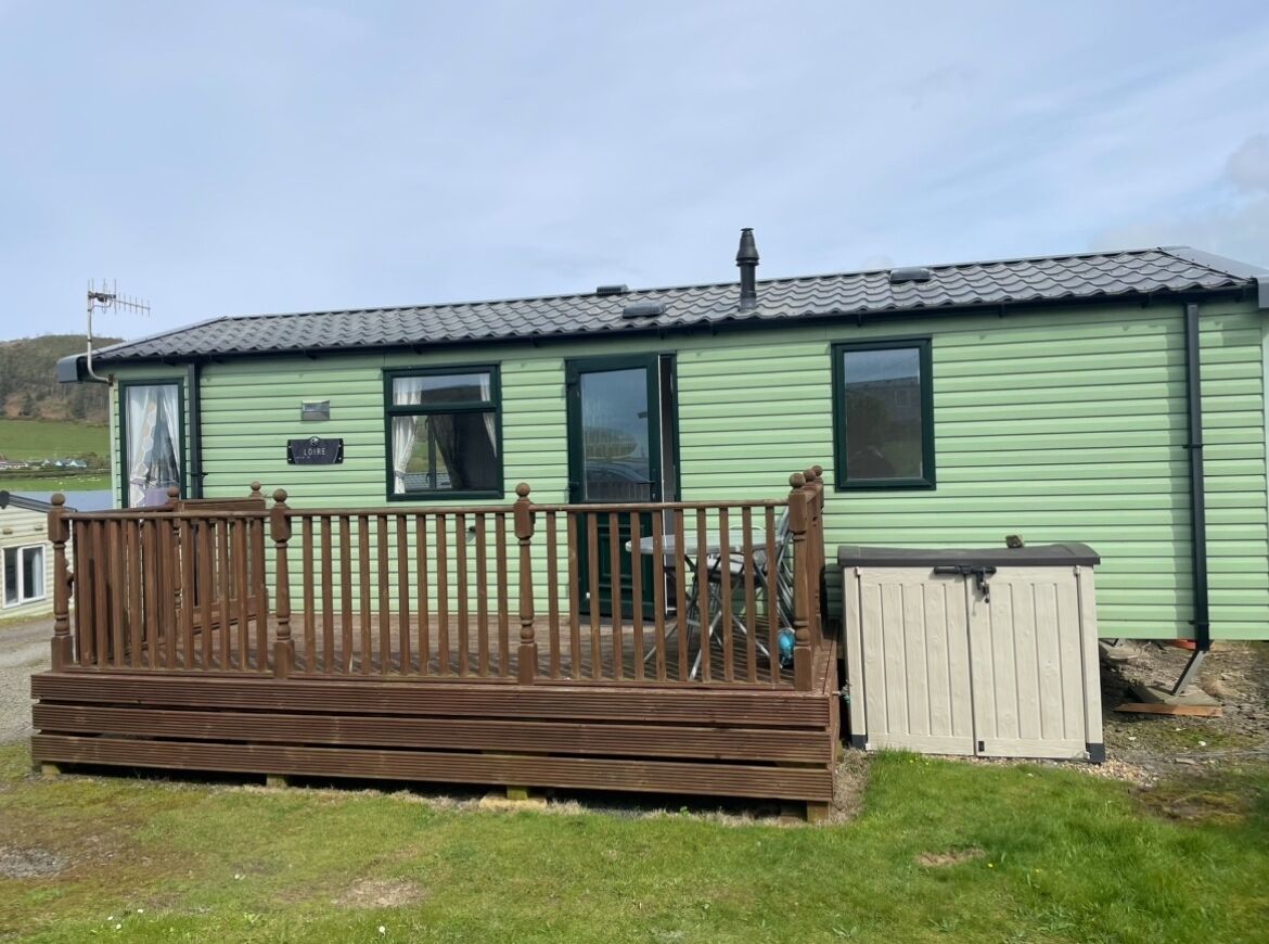 Une maison mobile verte avec une terrasse en bois et un petit abri de jardin.