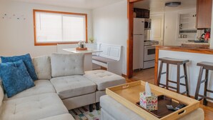 Una sala moderna con un sofá seccional, una mesa de centro de madera y una cocina visible a través de un vano abierto.