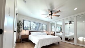 Une chambre moderne avec un grand lit, un plancher en bois, un ventilateur de plafond et une grande fenêtre équipée de stores.