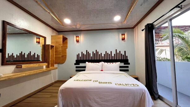 A modern hotel room with a large bed, a cityscape mirror, wall-mounted lights, and a view of palm trees. - swims maldives (Omadhoo)