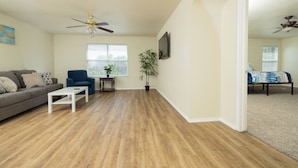 A living room with hardwood flooring, a gray sofa, a blue armchair, a white coffee table, a ceiling fan, a potted plant, and a TV mounted on the wall. - This home is minutes away from:
SEA WORLD
FIESTA TEXAS
RIVERWALK
4 JOINT BASES + (San Antonio)