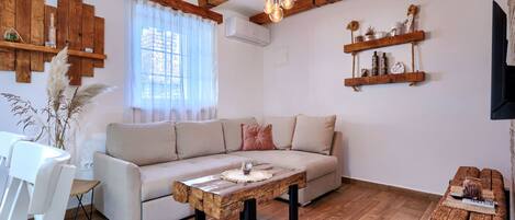 A living room with a sofa, wooden coffee table, and a TV. There are wooden beams on the ceiling and a window with sheer curtains.