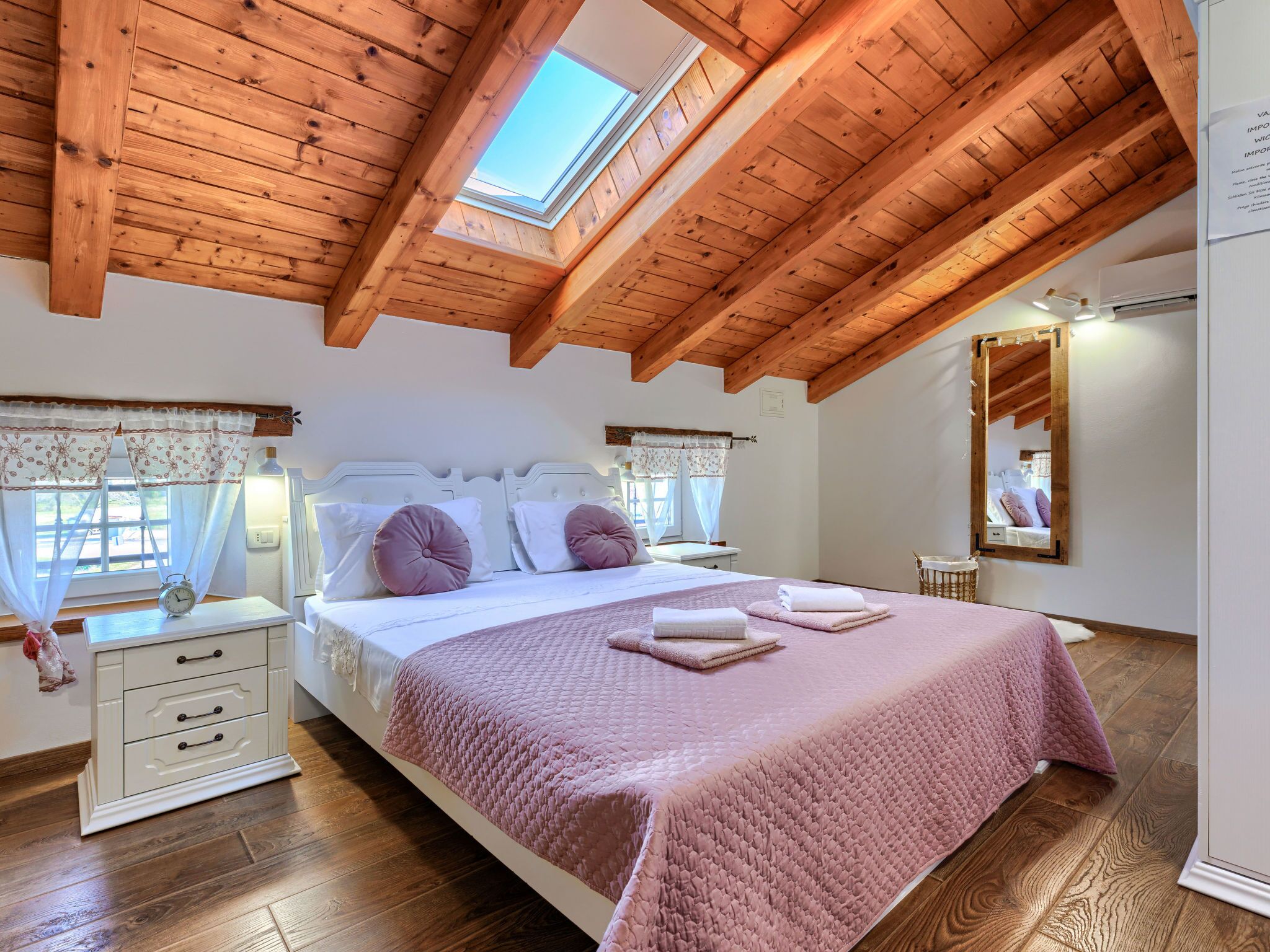 A bedroom with a sloped wooden ceiling, a skylight, a bed with pink bedding, a bedside table with a clock, and a mirror on the wall.