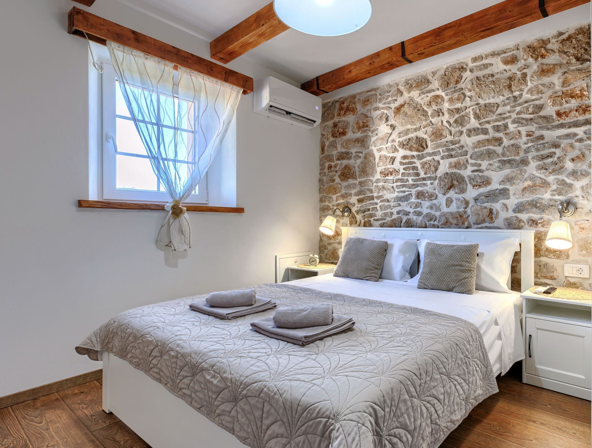 A bedroom with a stone wall, a bed with a quilt, two bedside lamps, and a window with a curtain.