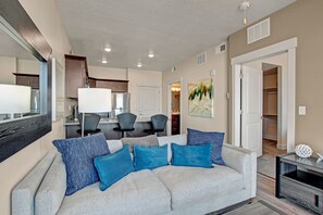 Een moderne woonkamer met een grijze bank, blauwe kussens, een eethoek met tafel en stoelen, en een keuken op de achtergrond.