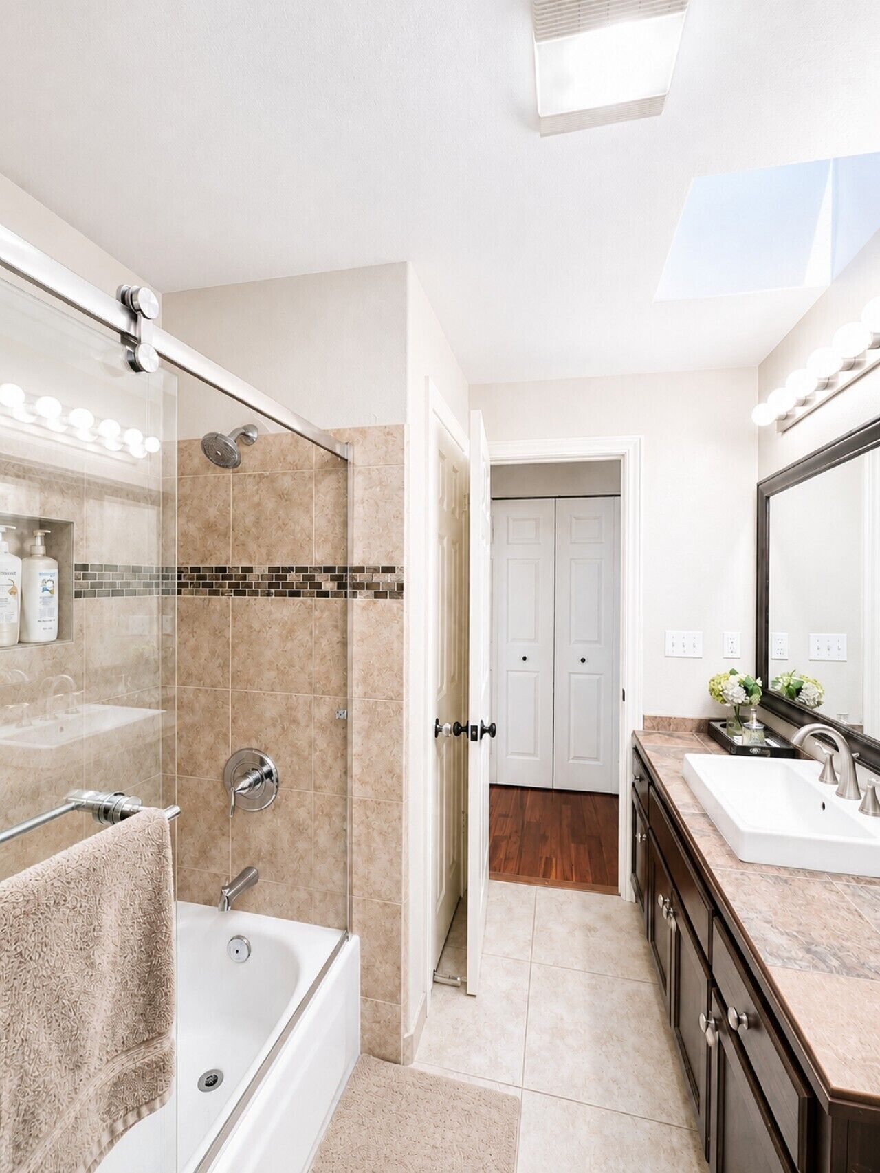 Une salle de bain moderne avec une douche à l’italienne en verre, une baignoire, un meuble-lavabo avec un miroir et une porte donnant sur une autre pièce.