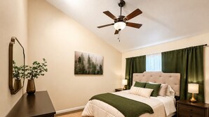 A bedroom with a bed, a ceiling fan, a nightstand, a dresser, a mirror, a painting, and a window with curtains.