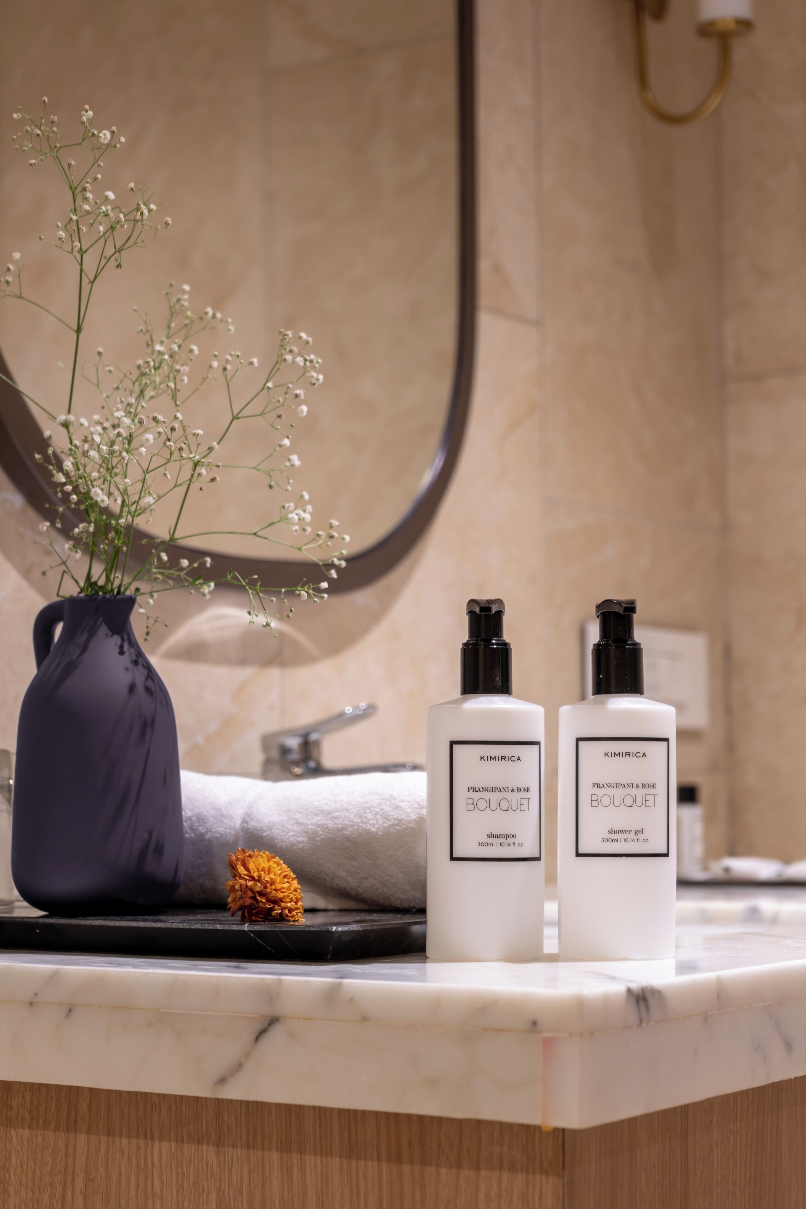 Un meuble-lavabo de salle de bain avec un vase de fleurs, deux bouteilles de shampoing et une serviette.