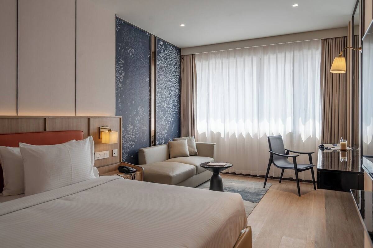 Quarto de hotel com uma cama grande, um sofá, uma escrivaninha e uma cadeira. Há uma janela com cortinas e uma luminária sobre a escrivaninha.