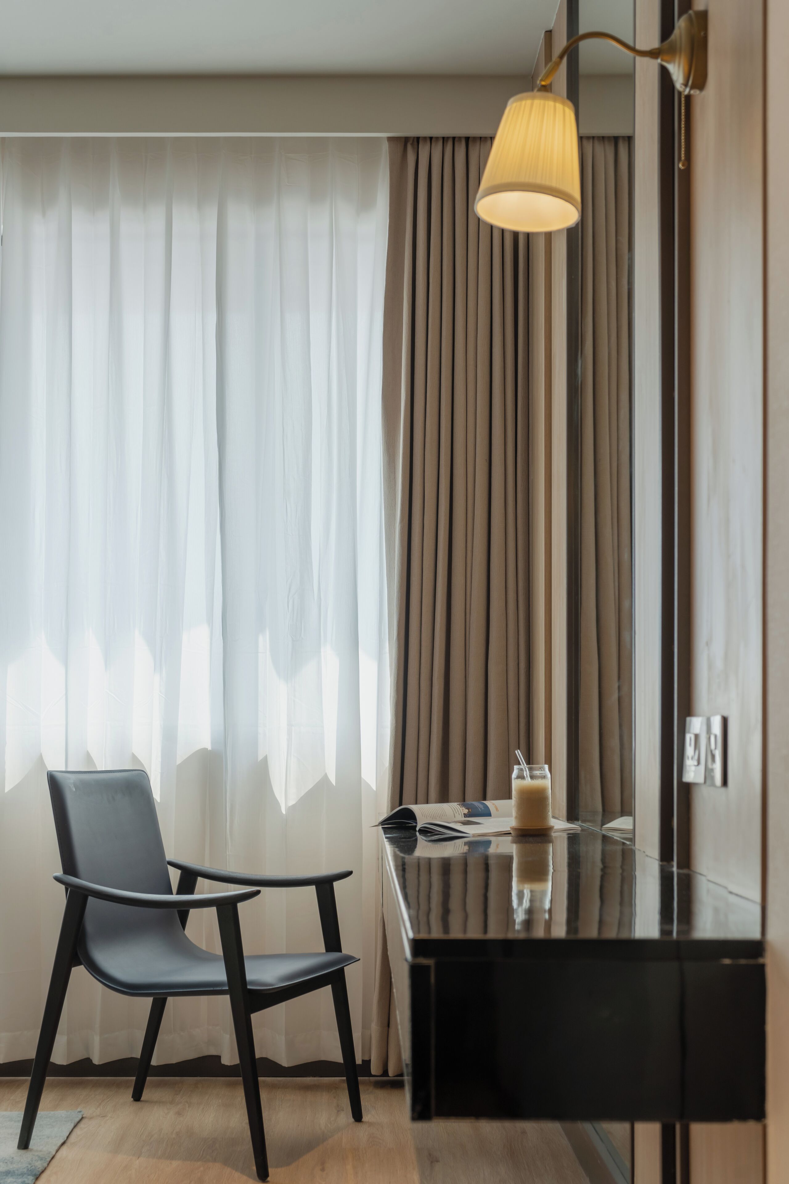 A modern hotel room with a black desk, a chair, a lamp, and a window with curtains.