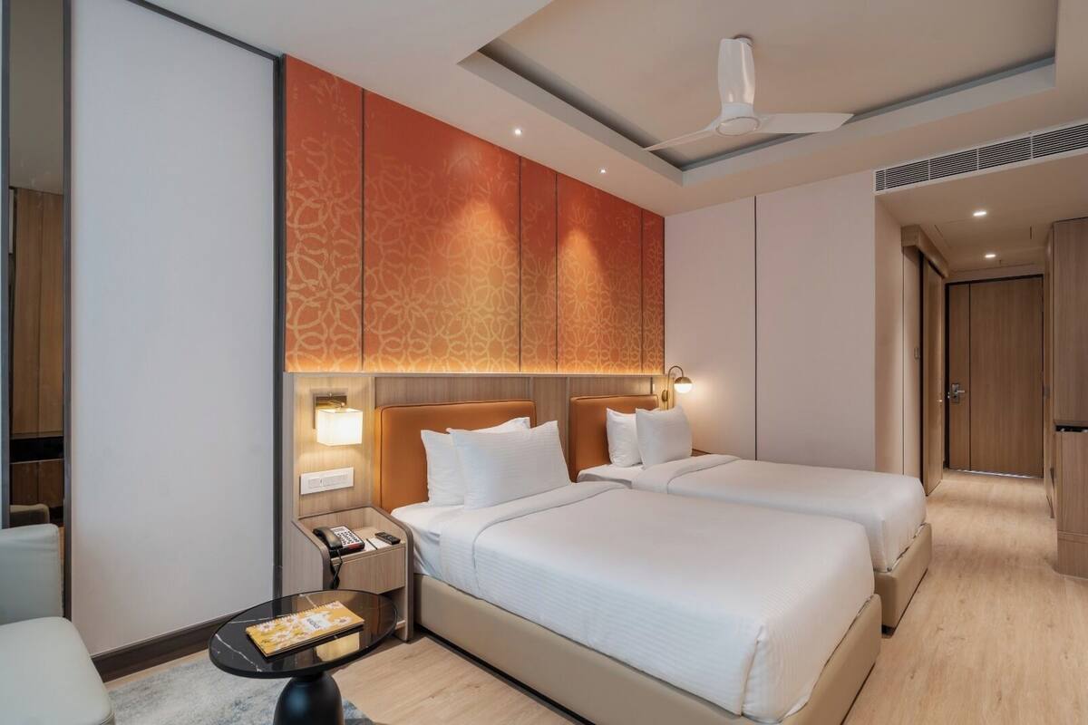 Quarto de hotel com duas camas, uma mesa de cabeceira com abajur, uma mesinha com um livro e um ventilador de teto.