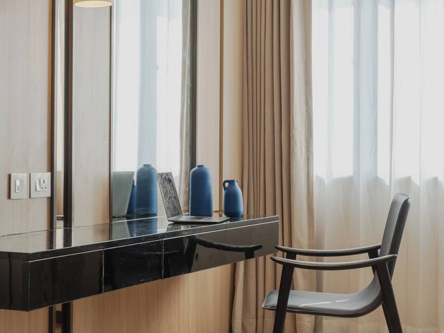 Una habitación de hotel moderna con un tocador negro, una silla, un espejo grande y una ventana con cortinas traslúcidas.