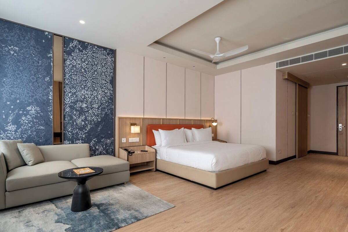 Quarto de hotel moderno com cama, sofá, mesa de cabeceira e um grande painel de parede abstrato.