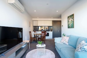 A modern living room with a flat-screen TV, a round coffee table with a plant, a light blue sofa, and a kitchen in the background.