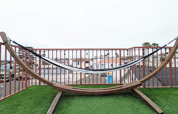A hammock on a grassy platform with a metal railing, overlooking a residential area with houses and parked cars.