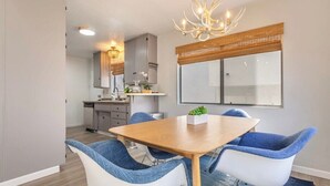 A modern dining area with a wooden table and blue and white chairs, a chandelier, and a large window with a view. - 3BR Townhouse in Prime Manhattan Beach Location (Manhattan Beach)