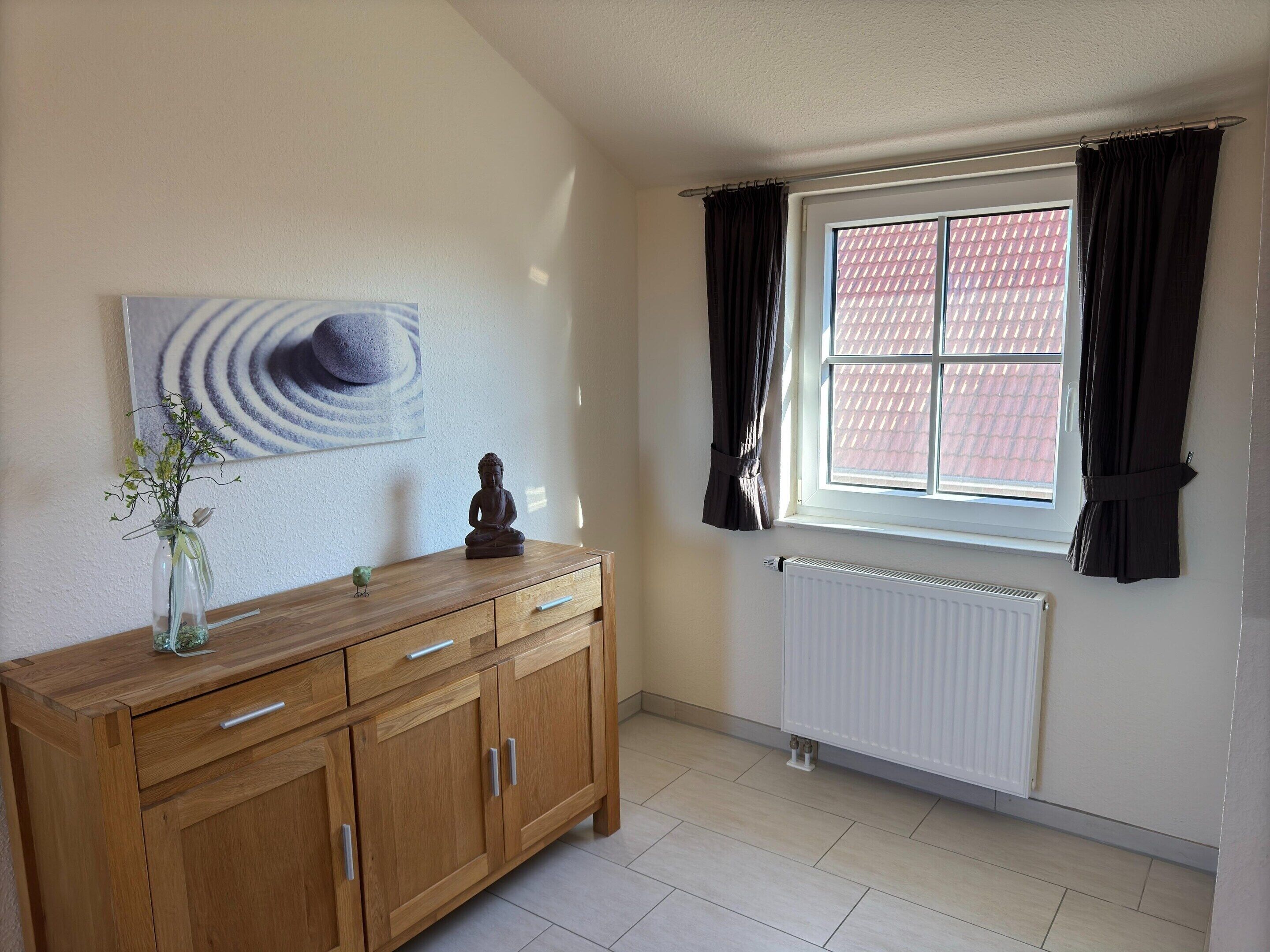 A room with a wooden cabinet, a Buddha statue, a vase with flowers, a painting, and a window with curtains.