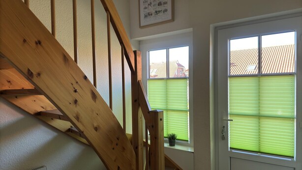 A wooden staircase leading to a landing with a radiator below. There are two windows with blinds, one of which has a plant on the sill. - Doppelhaushalfte in Burhave (Butjadingen)