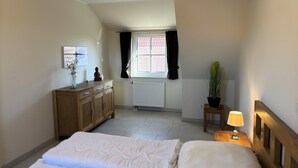 A hotel room with a bed, a wooden dresser, a radiator, a window with curtains, and a bedside lamp. - Doppelhaushalfte in Burhave (Butjadingen)