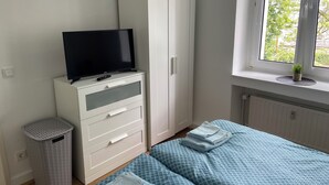 A bedroom with a bed, a TV on a dresser, a wardrobe, a window with a view of greenery, and a radiator. - Pempelforter Domicile (Düsseldorf)