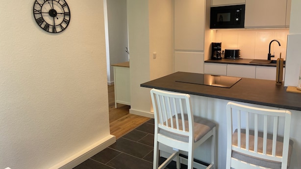 A modern kitchen with white cabinets, a black countertop, and a built-in microwave. There are two white chairs and a clock on the wall. - Pempelforter Domicile (Düsseldorf)