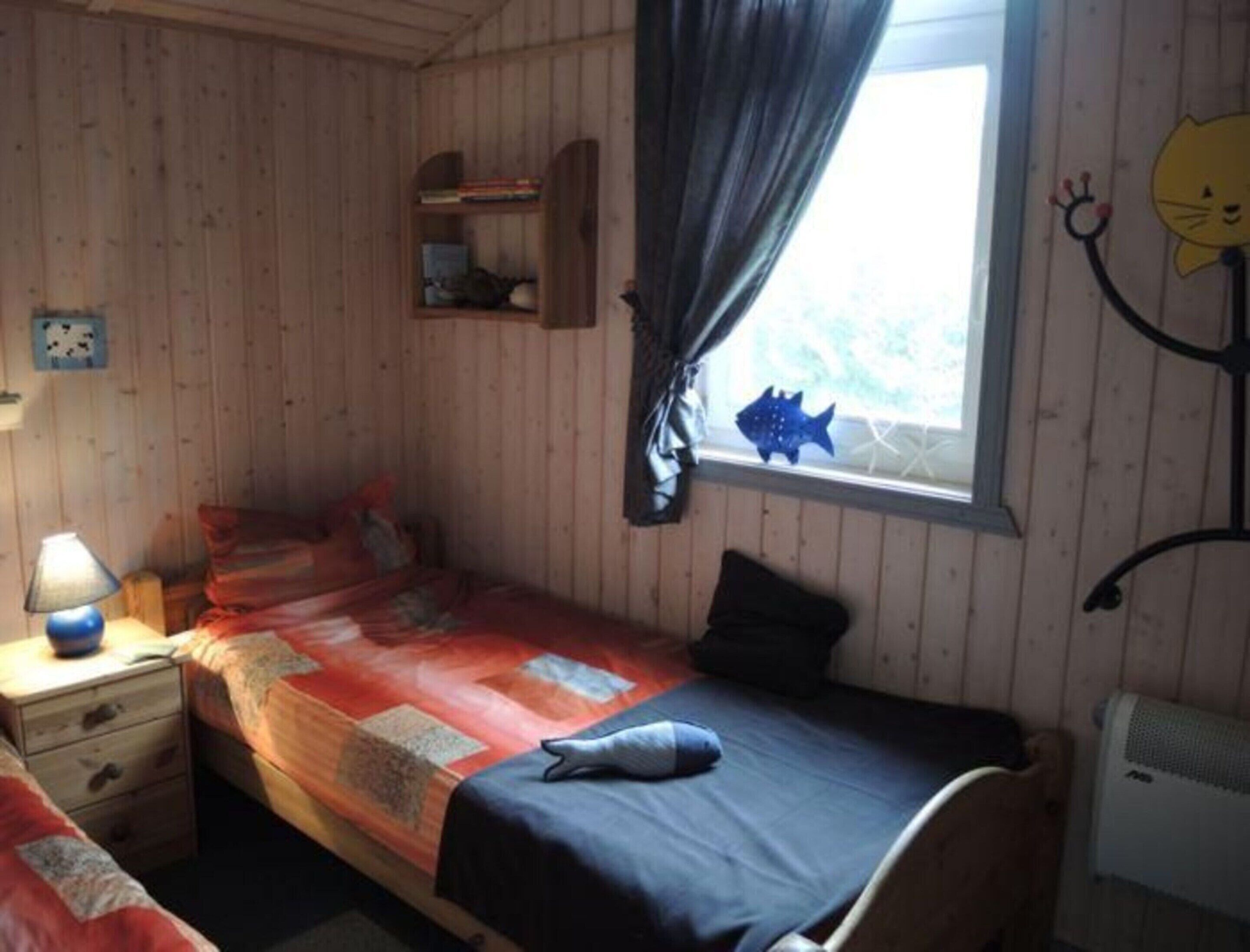Ein Einzelbett mit blauer Decke, ein Nachttisch mit Lampe, ein Regal mit Büchern und ein Fenster mit Vorhängen.